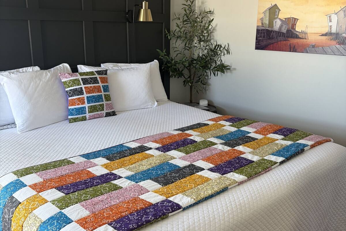 A bed with a colorful quilt, a dark headboard, a wall-mounted lamp, and a painting of a coastal scene.