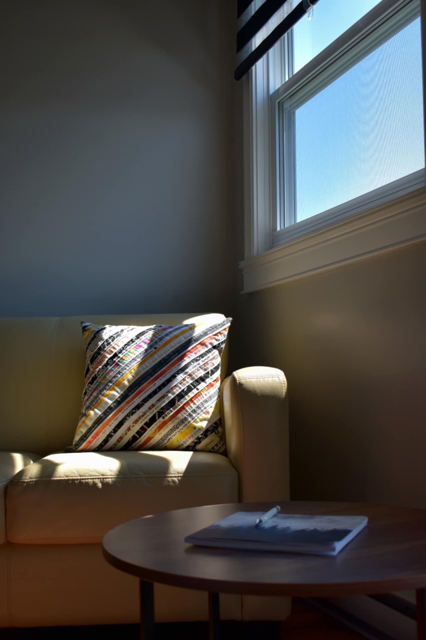 A corner of a room with a sofa, a patterned pillow, and a small round table with a book and pen.