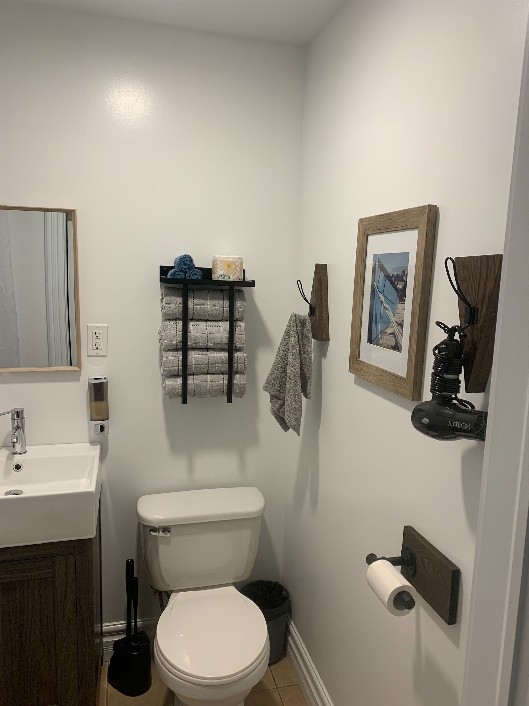A bathroom with a white sink, toilet, and a towel rack. There is a framed picture on the wall and a shelf with towels and toiletries.