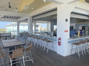 An outdoor bar area with tables and chairs, a counter with a person working behind it, and a view of buildings in the background. - Gorgeous Views! Oceanfront 4/4 Home - Pointe West access 38. Beachfront Bliss! (Galveston)