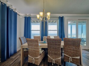 A dining area with wicker chairs, a chandelier, and large windows with blue curtains.