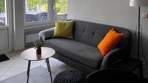 A modern living room with a grey sofa, a small round table, and a black ottoman. - Moderner Tiny Bungalow am See in der Oberlausitz, zum Radfahren, Wandern, Angeln (Quitzdorf am See)