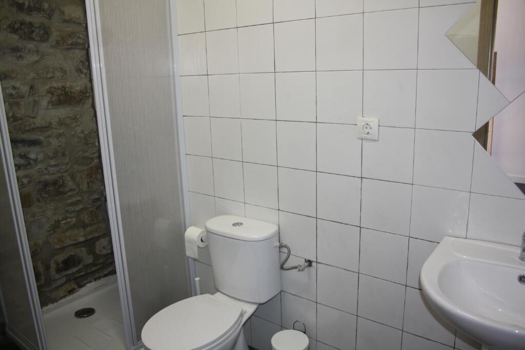 Een badkamer met witte betegelde wanden, een wit toilet, een witte wastafel en een gedeeltelijk zichtbare stenen muur.