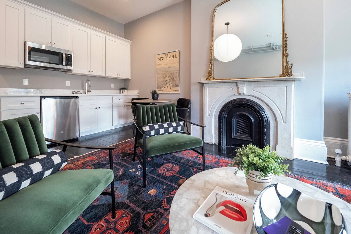 Una cocina moderna con armarios blancos, un refrigerador de acero inoxidable y un microondas. Se observa un comedor con una silla tapizada de color verde y un tapete estampado. Se puede ver una chimenea con un espejo enmarcado en dorado.