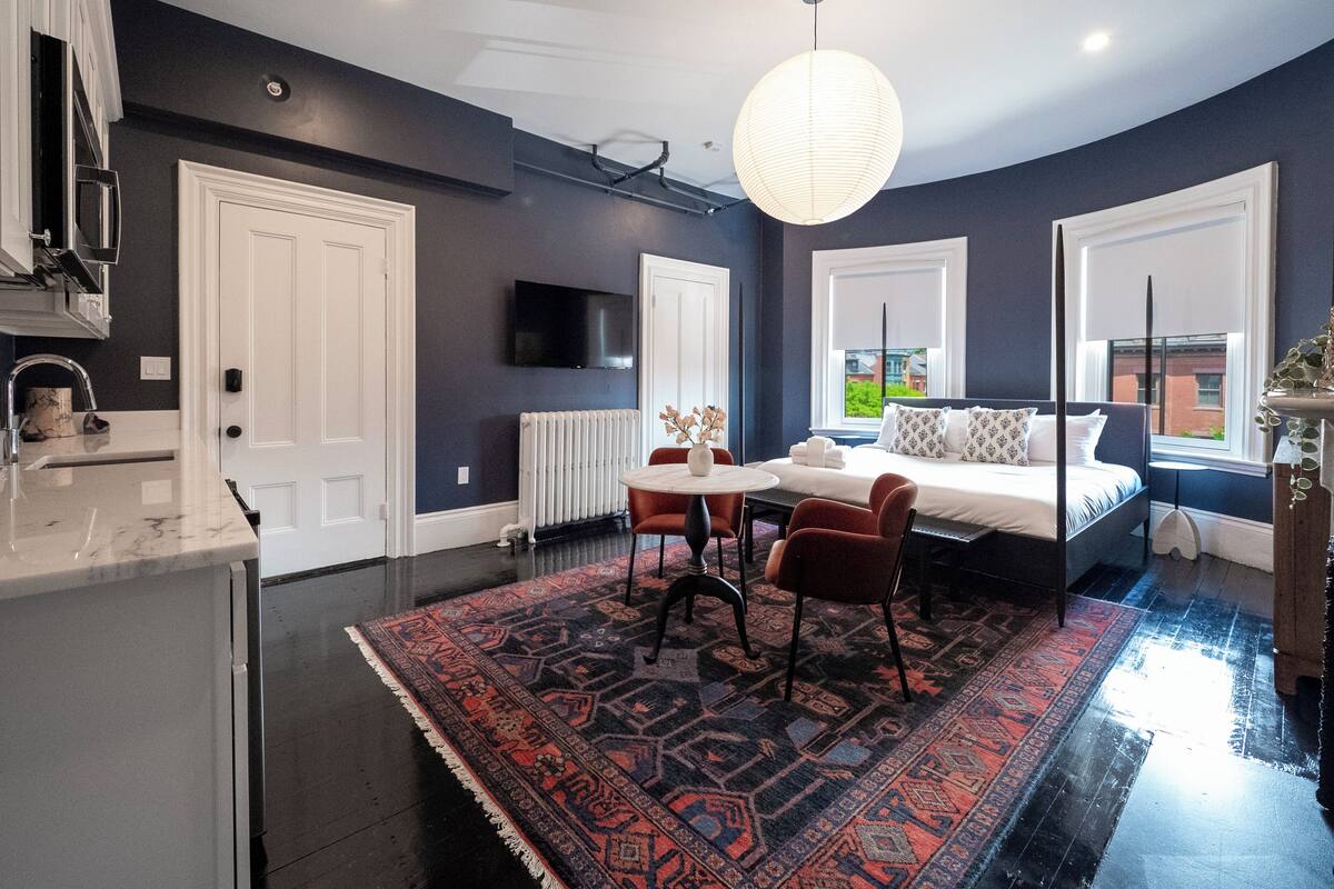 Una habitación de hotel moderna con una cama grande, un área de comedor con mesa y sillas, una cocina con un lavaplatos y alacenas, y un ventanal que ofrece vistas al exterior.