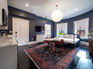 A modern hotel room with a large bed, a dining area with a table and chairs, a kitchen with a sink and cabinets, and a large window providing a view of the outside. - The Chester Hotel (Boston)