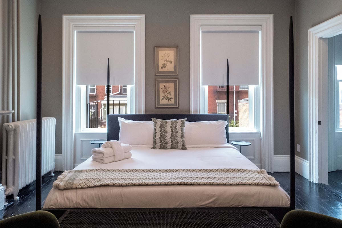 Un dormitorio con una cama grande, dos ventanas, un radiador y un cuadro enmarcado en la pared.