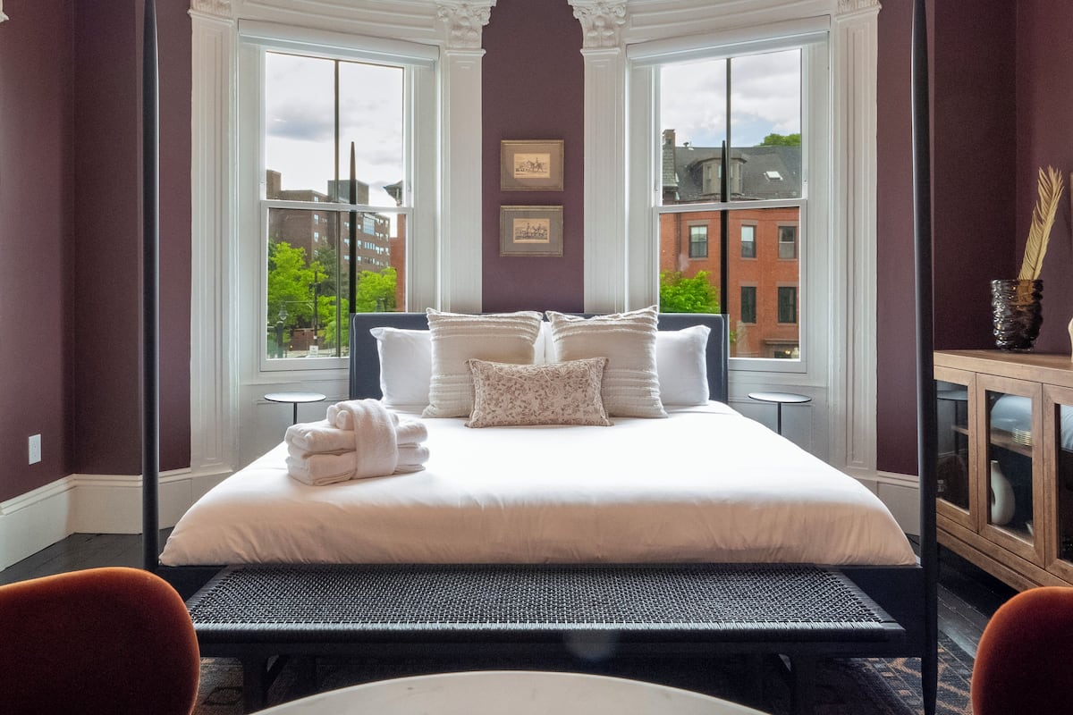 Habitación de hotel con una cama grande, dos ventanas, vistas a edificios y un mueble con un florero.
