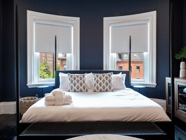 Habitación de hotel con una cama grande, dos ventanas con persianas, vista a edificios y una mesita con un florero.