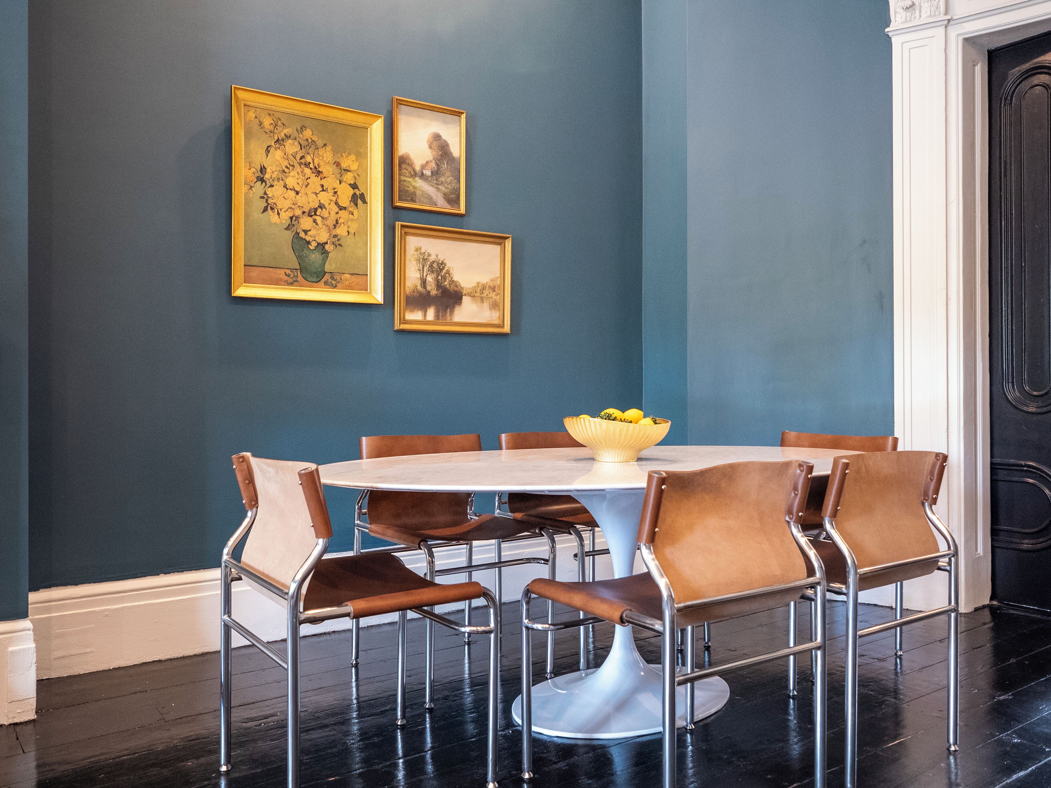Un coin repas avec une table ronde, six chaises et un saladier rempli de fruits. Trois tableaux encadrés ornent le mur.