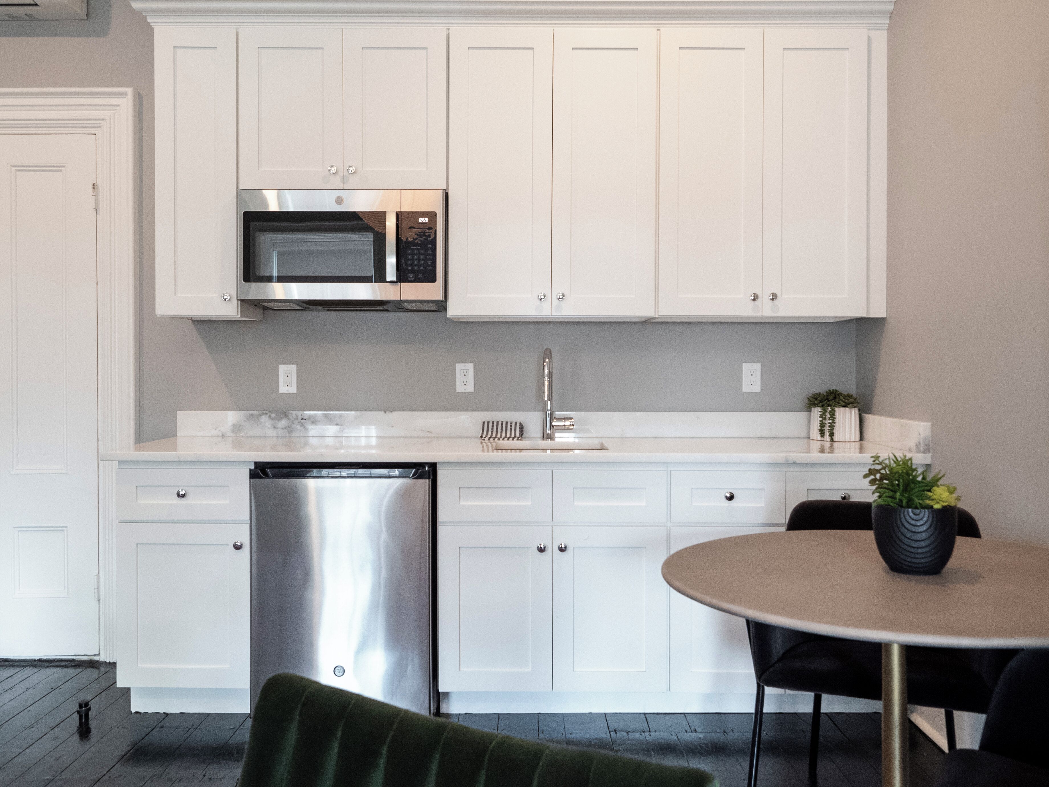 A modern kitchen with white cabinetry, a stainless steel dishwasher, a built-in microwave, and a round dining table with a potted plant.