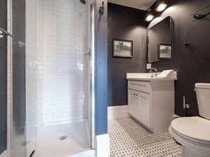 A modern bathroom with a glass shower enclosure, white tiled walls, a dark accent wall, a white sink with a mirror above it, and a toilet. - The Chester Hotel (Boston)
