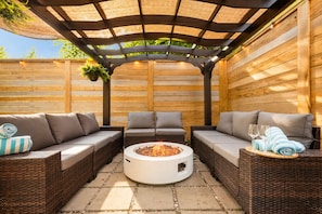A covered outdoor seating area with wicker furniture, a central fire pit, and a pergola overhead.