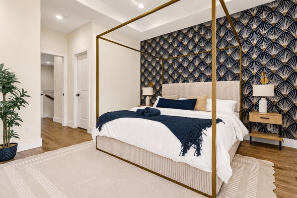 A bedroom with a four-poster bed, a patterned wallpaper, a bedside table with a lamp, and a potted plant.