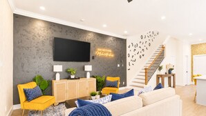 A modern living room with a flat-screen TV, a sofa, two yellow chairs, a staircase, and a wall with a neon sign.