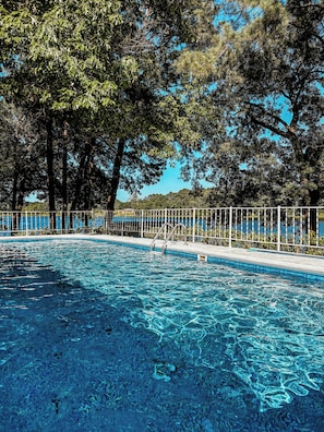 A clear swimming pool surrounded by a white fence, with trees and a clear blue sky in the background. - The Hap Haven- a Charming Lakefront Retreat in East Texas - Perfect Getaway! (Lone Star)