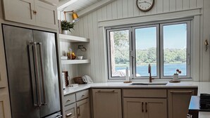 A modern kitchen with a stainless steel refrigerator, wooden cabinets, a window with a view of a lake, and a clock on the wall. -  The Hap Haven- a Charming Lakefront Retreat in East Texas - Perfect Getaway! (Lone Star)