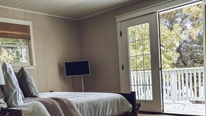 A bedroom with a bed, a desk, a chair, a television, and a balcony with a view of trees. -  The Hap Haven- a Charming Lakefront Retreat in East Texas - Perfect Getaway! (Lone Star)