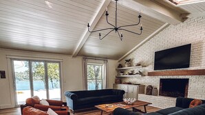 A spacious living room with a high ceiling, a fireplace, a television mounted on the wall, and a large window offering a view of the outdoors. -  The Hap Haven- a Charming Lakefront Retreat in East Texas - Perfect Getaway! (Lone Star)