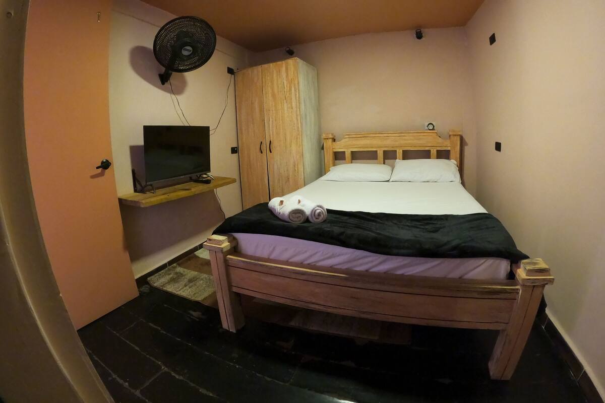 A bedroom with a wooden bed, a TV on a shelf, a wardrobe, and a fan on the wall.