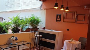 Un patio acogedor con un sofá marrón, una mesa de centro de vidrio y una barra con taburetes. Hay plantas en macetas e iluminación colgante.