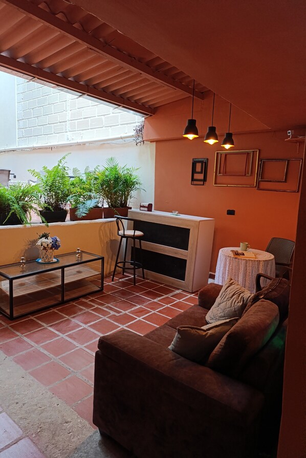 A cozy patio area with a brown sofa, a glass coffee table, and a bar area with stools. There are potted plants and hanging lights. - Casa Primavera Hostal (Medellín)