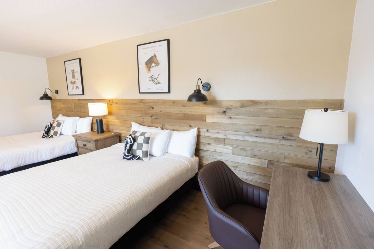 A hotel room with two beds, a wooden headboard, a desk with a chair, and wall-mounted lamps.