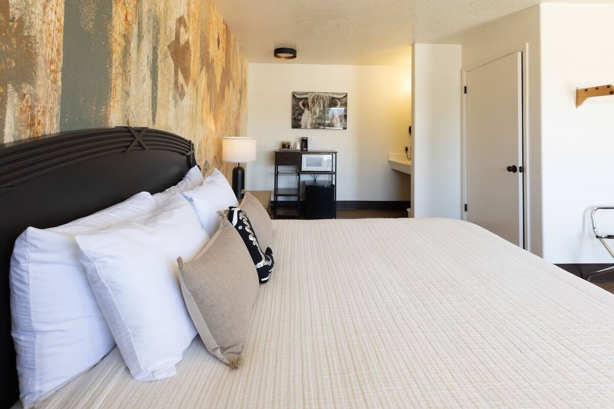 A hotel room with a large bed, a headboard, a nightstand, a lamp, a microwave, a small table, and a door.