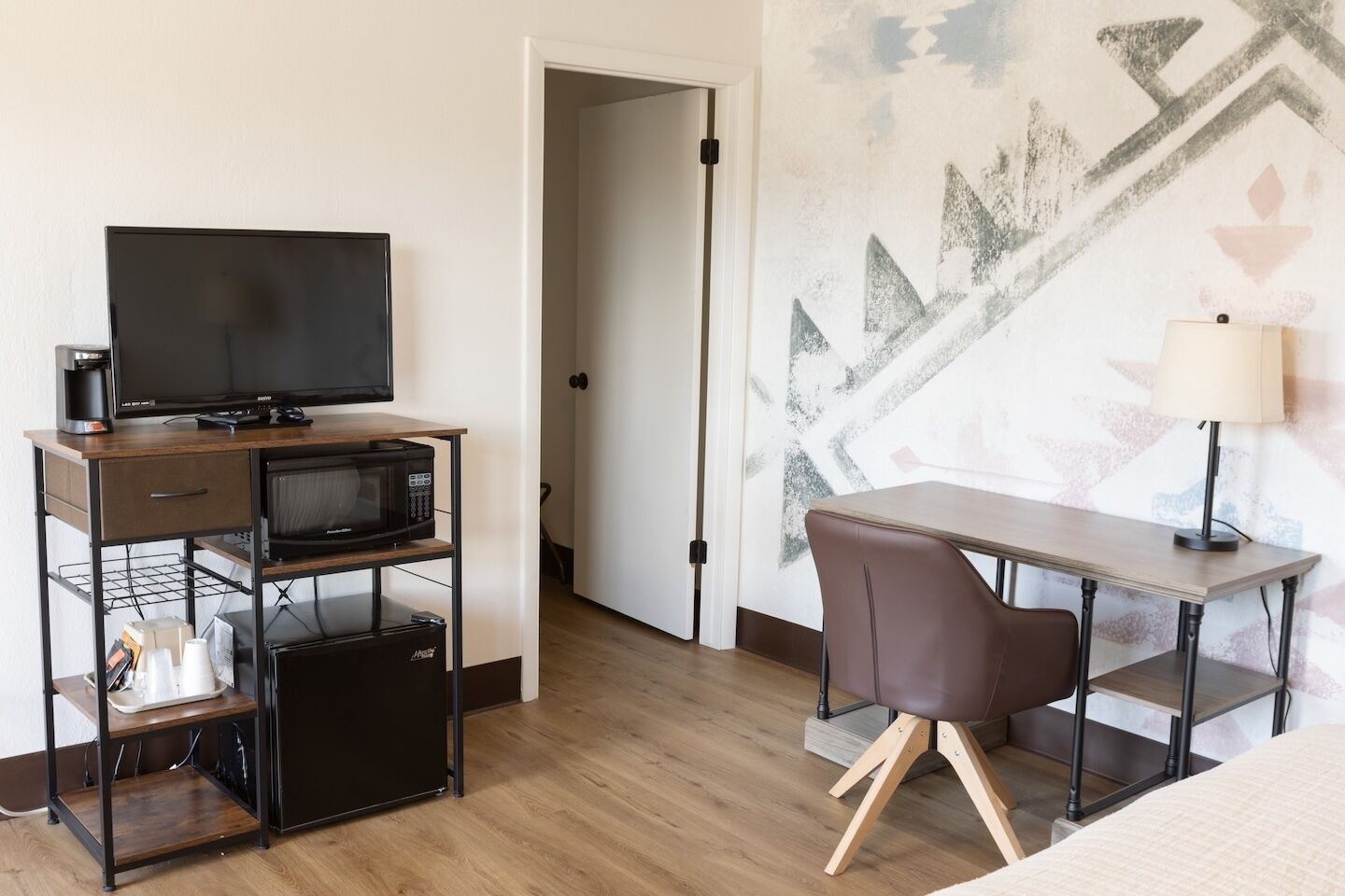Et lille hotelværelse med skrivebord i træ, fjernsyn, mikroovn, stol og seng.