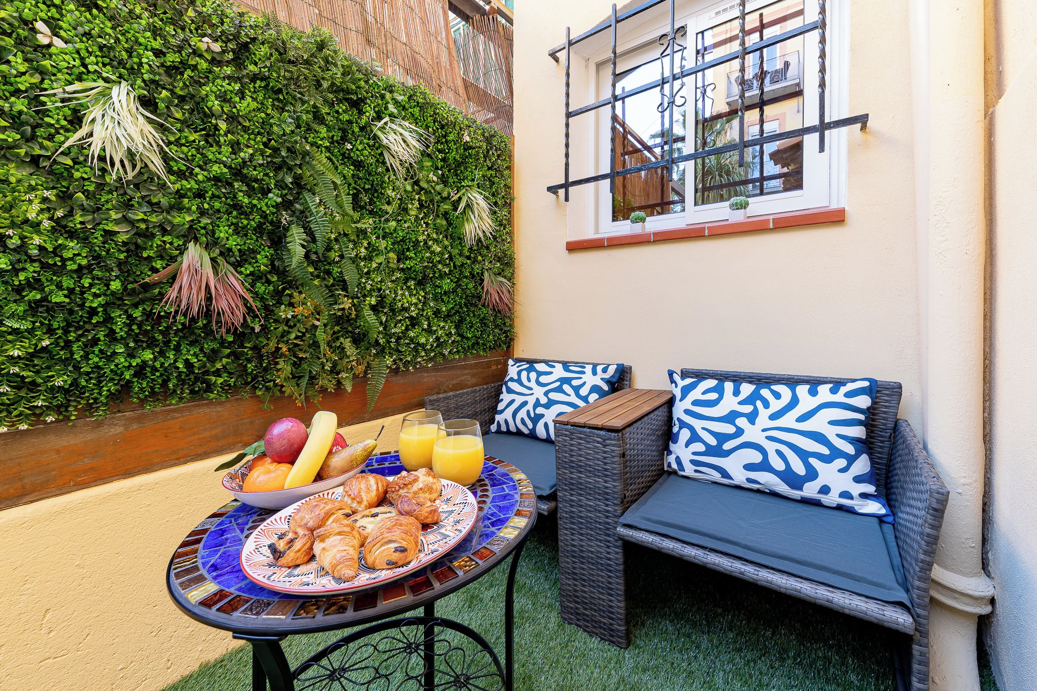Un espace extérieur aménagé avec une table dressée pour le petit-déjeuner, comprenant des croissants et des fruits, ainsi qu’un canapé en osier agrémenté de coussins à motifs.
