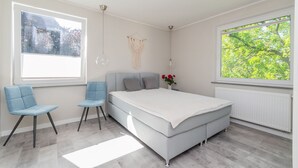 Ein Schlafzimmer mit einem Bett, zwei Stühlen, einer Wanddekoration aus Makramee und einem Fenster mit Blick auf Bäume.