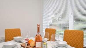 Una mesa de comedor dispuesta con platos, cuencos y vasos blancos, una botella de vino y una tabla de madera con frutas y una vela.