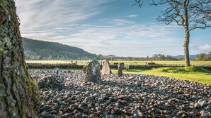 Un círculo de piedras con una cruz al fondo, rodeado por un campo y una valla.