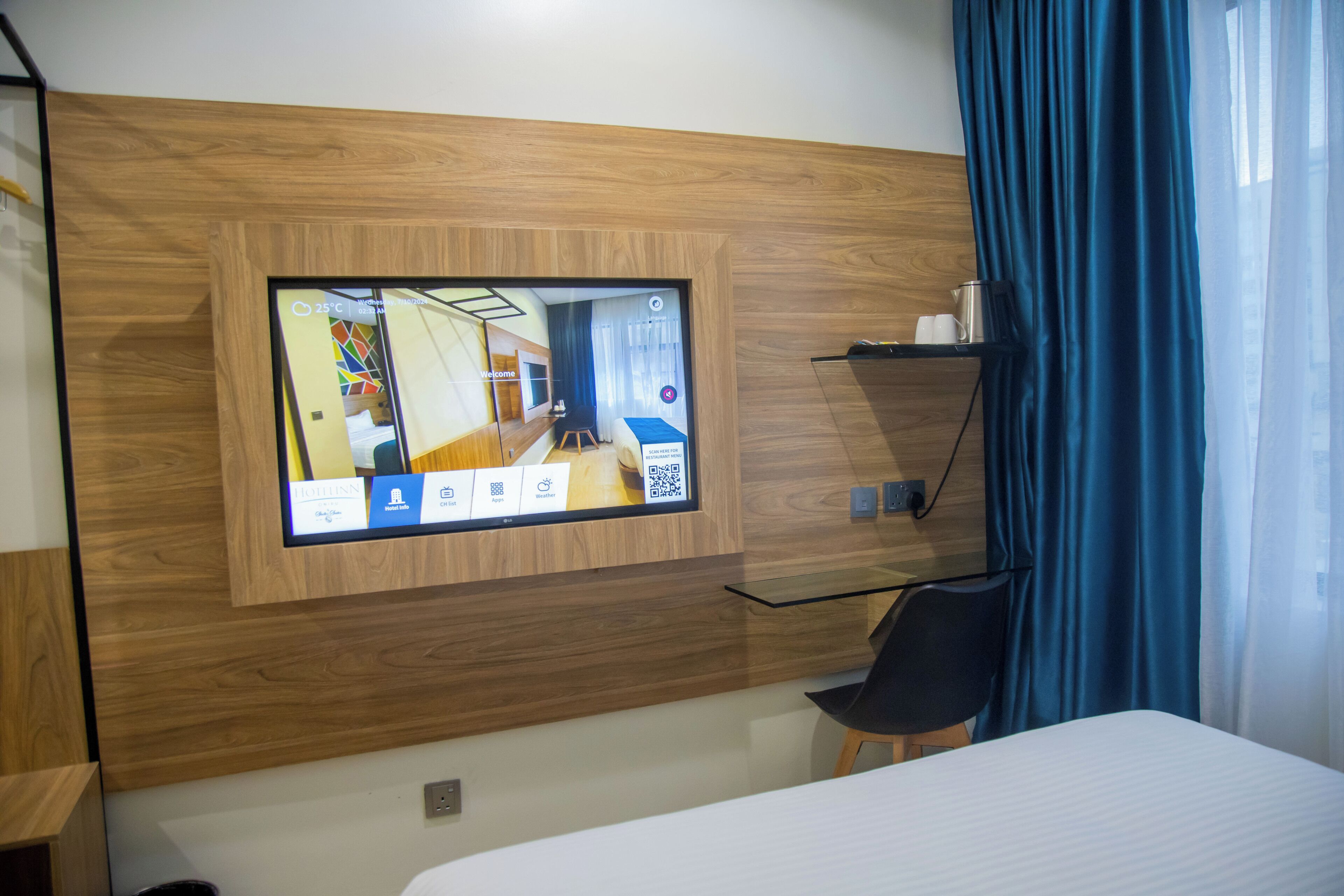 Camera d'albergo con TV a parete che mostra una planimetria, un letto, una scrivania, una sedia e uno scaffale con oggetti.