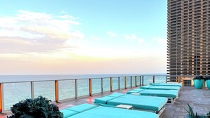 Een terras op het dak met meerdere ligbedden, uitzicht op zee en een hoog gebouw op de achtergrond.