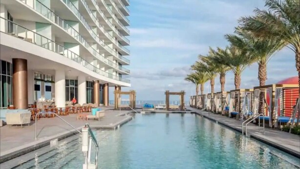 Een modern hotel met een groot zwembad, terras en palmbomen.