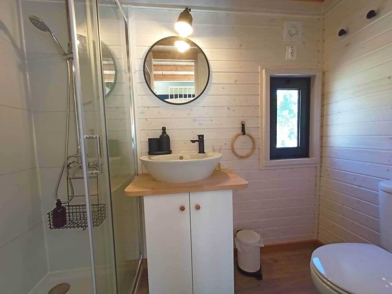 Een badkamer met een douchecabine van glas, een wit wastafelmeubel met een houten blad, een ronde spiegel en een klein raam.