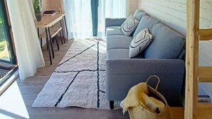 A cozy cabin interior with a wooden ceiling, a gray sofa with cushions, a dining table, and a window with curtains.