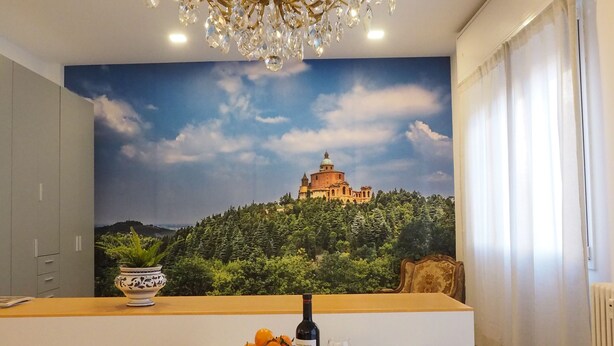 Ein Essbereich mit einem Glastisch, weißen Stühlen, einer Chandelüster und einem Wandbild eines Schlosses auf einer Anhöhe.