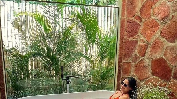 Une femme dans une baignoire, avec vue sur une végétation luxuriante à l’extérieur.