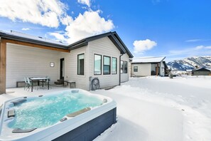 Ein Haus mit Whirlpool im Schnee, eine Terrasse mit Stühlen und Berge im Hintergrund.