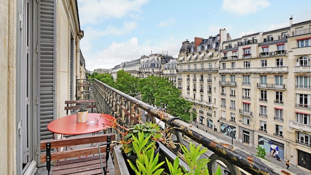 Un balcon avec une table et des chaises, donnant sur une rue de la ville, avec des immeubles et des arbres.