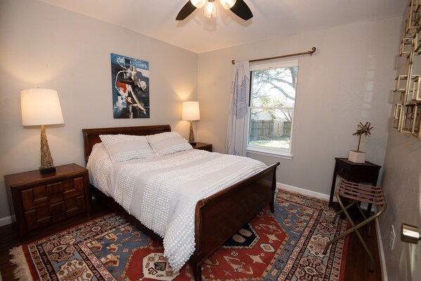 A bedroom with a wooden bed, two bedside lamps, a patterned rug, a window with curtains, and a wall-mounted shelf.