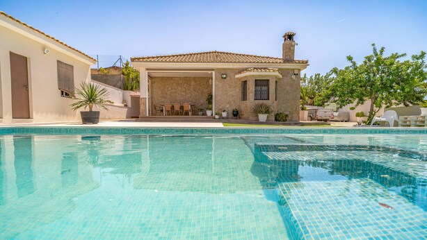 Une piscine avec une terrasse carrelée, un bâtiment en pierre avec un toit en tuiles et des plantes en pot.