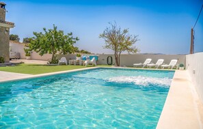 A rectangular swimming pool with clear blue water, surrounded by a white wall and a grassy area with white lounge chairs.