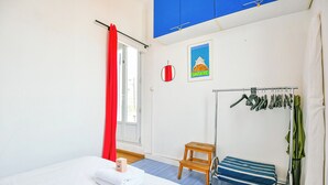 Et hotelværelse med en seng, en træskammel, et tørrestativ og et billede i ramme på væggen.