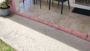 Ein überdachter Patio-Bereich mit zwei Korbstühlen, einem kleinen Tisch, einer Topfpflanze und einem Fenster mit Blick ins Freie.