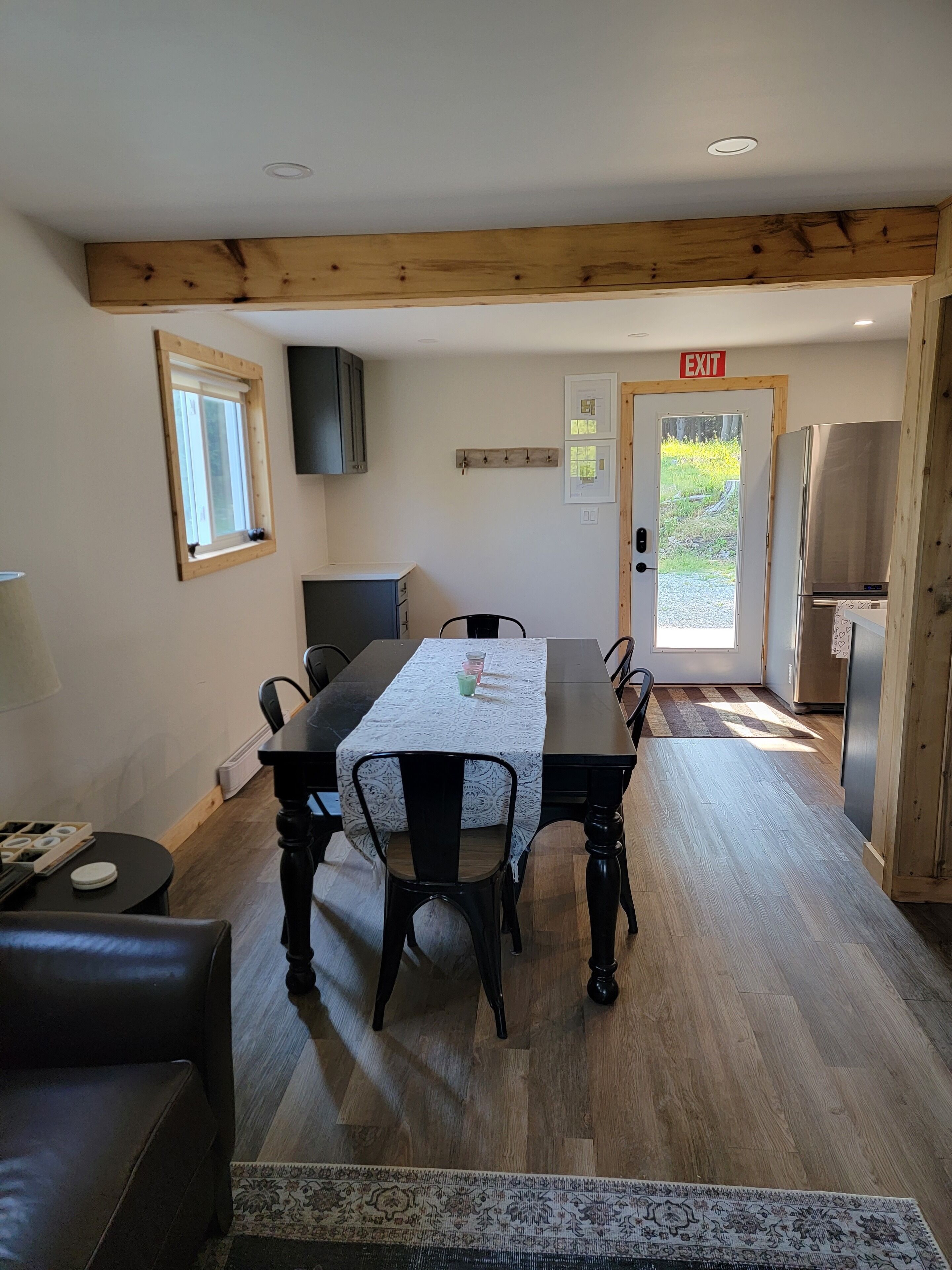 Un comedor con una mesa de madera, sillas, una mesita auxiliar y un sofá. Se ve una ventana, una puerta con letrero de salida y un refrigerador al fondo.