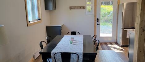 Un comedor con una mesa de madera, sillas, una mesita auxiliar y un sofá. Se ve una ventana, una puerta con letrero de salida y un refrigerador al fondo.
