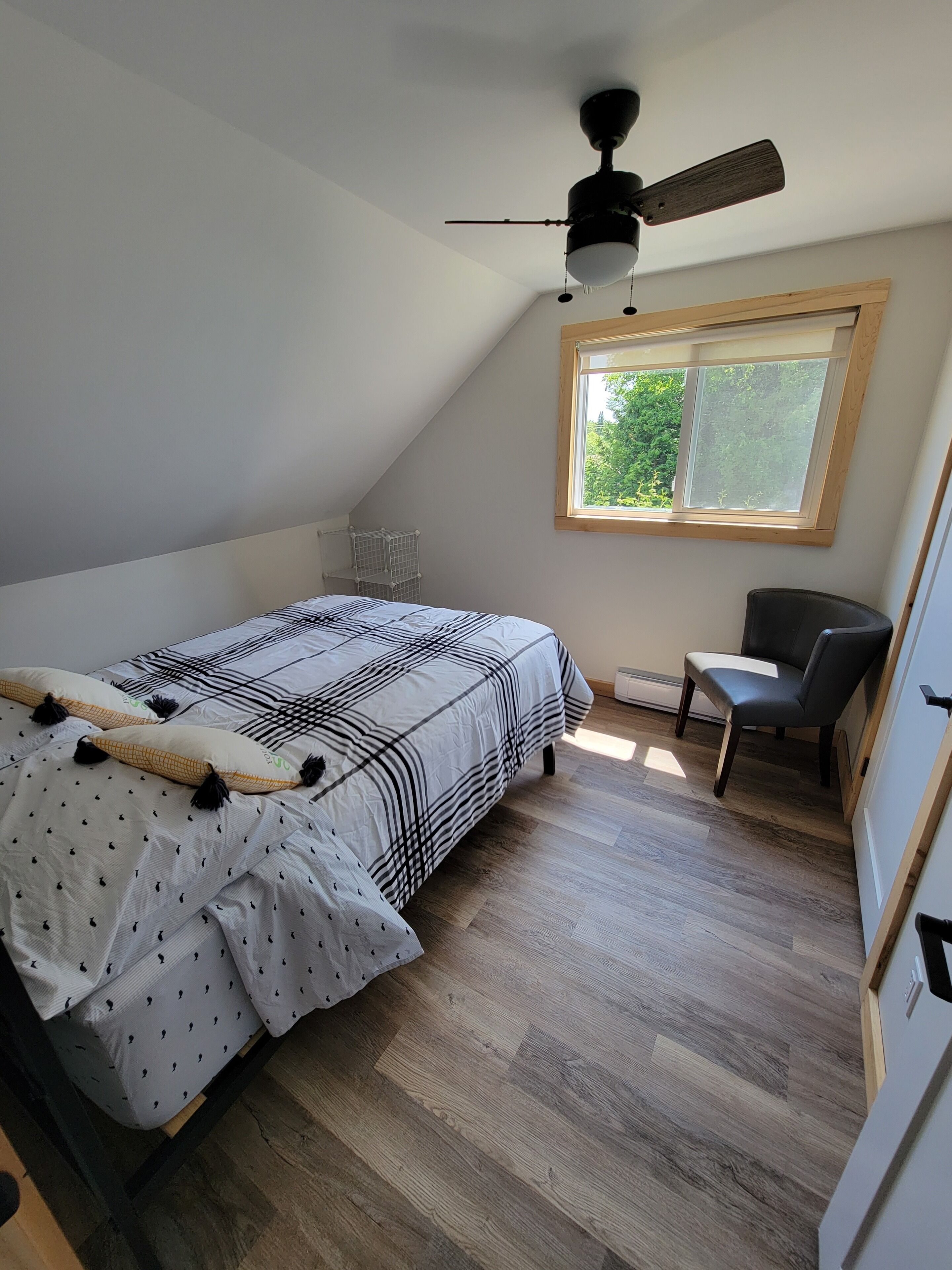 A bedroom with a bed, a desk, a chair, a ceiling fan, and a window with a view of greenery.